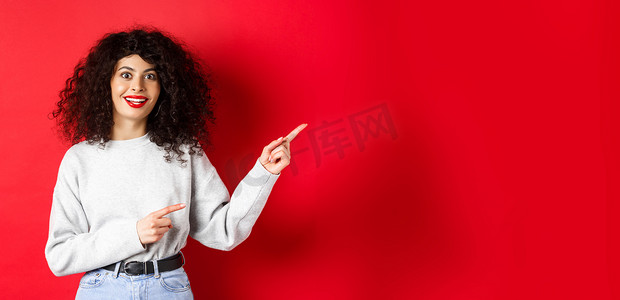 促销活动扁平风摄影照片_留着卷发的有趣的年轻女性，用手指指着标志，看起来印象深刻和兴奋，正在查看促销活动，红色背景