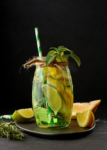 透明玻璃杯中的柠檬水，黑色背景中配有柠檬、酸橙、迷迭香小枝和薄荷叶