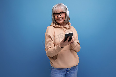 60 多岁的现代祖母，头发灰白，手里拿着智能手机，正在工作室背景和复制空间中研究小工具和耳机