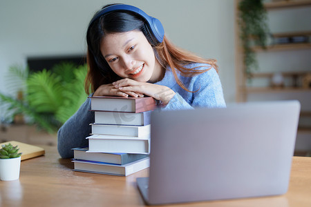 年轻美丽的亚洲女性在清晨在线课堂上以书籍、耳机和电脑作为家里的学习材料时露出笑脸的肖像