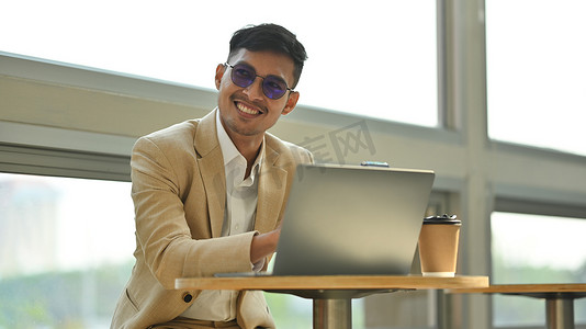 微笑的年轻商人在现代咖啡店用笔记本电脑工作的光致变色眼镜