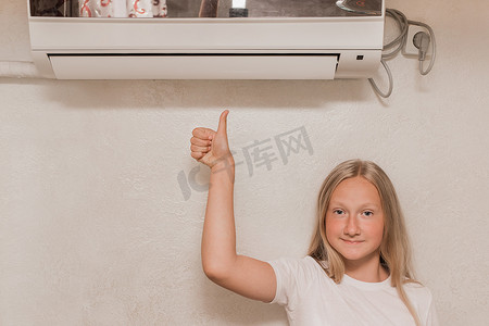 少女金发摄影照片_年轻可爱的少女金发欧洲人的外表显示出酷酷的拇指，看着镜头，在房间墙上的空调旁微笑