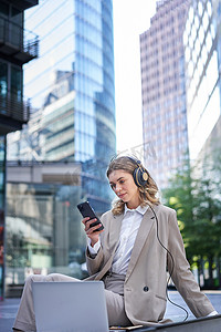 app新闻滚动摄影照片_穿着西装的企业女性，坐在城市里，用笔记本电脑听音乐，在手机上滚动新闻，午休时放松