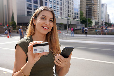 巴西圣保罗市保利斯塔大道金融中心手持智能手机并出示信用卡看着相机的微笑女性肖像