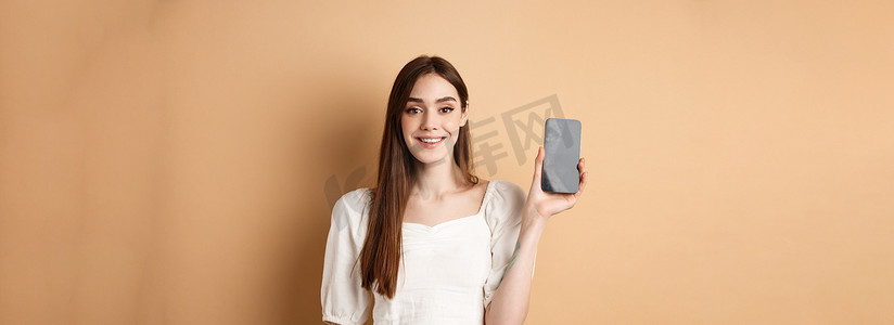 app展示摄影照片_快乐的女孩展示着空荡荡的手机屏幕，微笑着展示手机应用程序，站在米色背景中