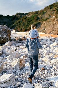 背大石头摄影照片_爸爸肩上扛着一个小女孩，沿着鹅卵石滩走向青山和大石头。
