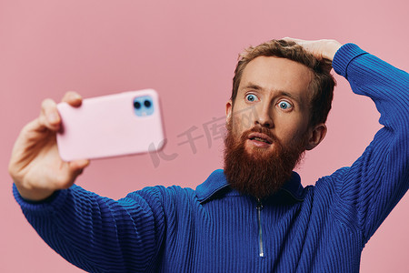 一位手持手机的红头发男子的肖像，他在手机上自拍和拍照，在粉红色背景下微笑，博主