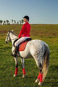 女骑手摄影照片_年轻的女骑手，穿着红色的红色长裤和白色的马裤，在夕阳的余晖中骑着她的马。