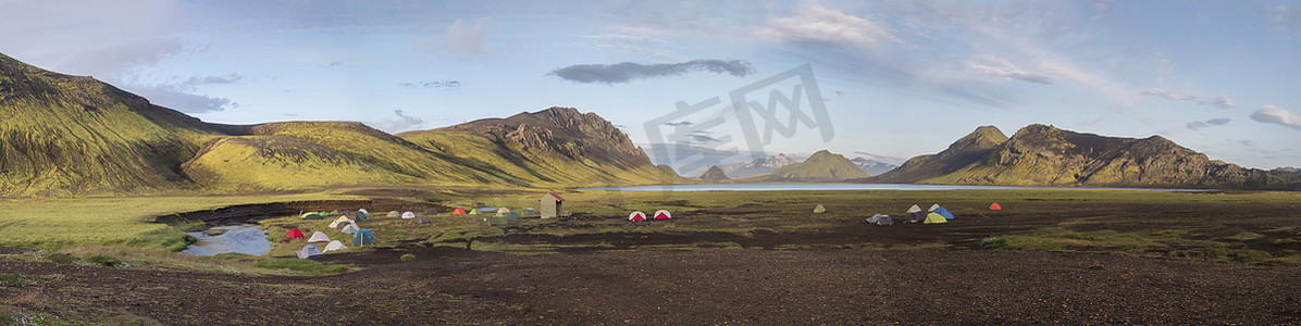 山丘背景摄影照片_全景景观与五颜六色的帐篷在露营地在蓝色的 Alftavatn 湖与绿色的山丘和冰川在 Fjallabak 自然保护区的美丽风景在 Laugavegur 远足径的冰岛高地部分。