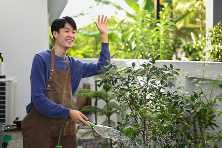 亚洲男性农民在后院浇水，用友好的笑脸问候邻居