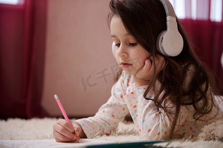 特写才华横溢的小女孩戴着耳机听音乐，用铅笔画画，躺在卧室的地毯上