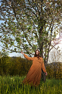 一位留着长长的红头发的苗条迷人的女人穿着橙色的长裙站在一棵开花的树旁的乡下，手里拿着裙子快乐地旋转着。