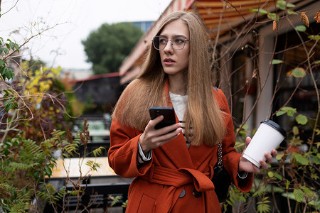年轻时尚的女商人手里拿着一杯咖啡，街上拿着手机，穿着橙色的秋装，经营着一家成功的商业概念