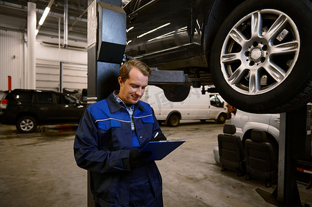 汽车机械师、技术员、身穿制服的汽车工程师在剪贴板上制作清单，用于在保修汽车维修期间修理在维修车间用起重机吊起的汽车。