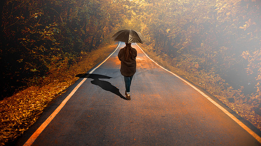 一个身穿黑色衬衫和蓝色牛仔裤的年轻女孩手里拿着一把黑色雨伞走在路上的照片。