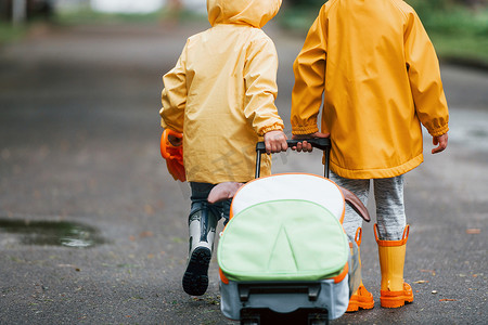 防水雨伞摄影照片_两个带着雨伞、手提箱和黄色防水斗篷和靴子的孩子在雨后一起在户外散步