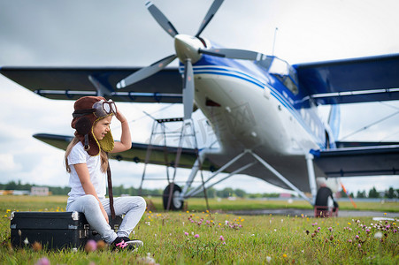 一个穿着飞行员服装的小女孩坐在机场的复古手提箱上等待航班起飞。