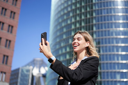 挥手送客摄影照片_在街上微笑的企业女性视频通话的肖像，手持手机，对着智能手机摄像头挥手，穿着西装