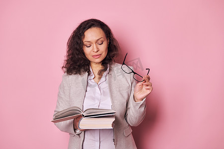 一头卷发的黑发成年女性手里拿着眼镜，在粉红色背景下阅读学习书