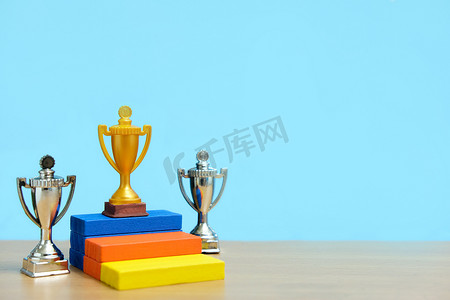金属奖杯摄影照片_金色奖杯站在色彩缤纷的领奖台上，两座银色奖杯