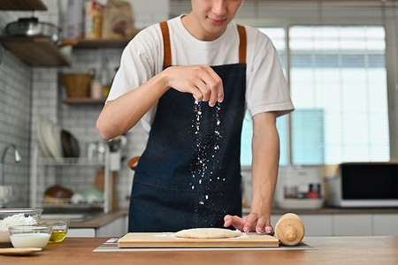 年轻人穿着围裙在新鲜面团上撒面粉，同时在餐桌上准备自制糕点