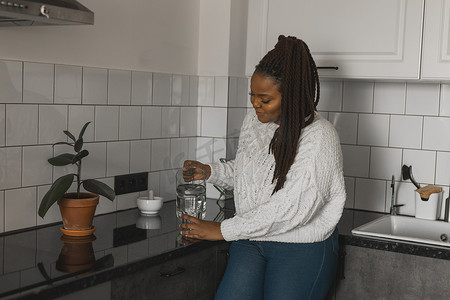 喝矿泉水摄影照片_口渴的非洲裔美国妇女在厨房里喝矿泉水，从水壶里倒健康的液体到玻璃杯里，带辫子的快乐女士享受清爽的饮料，复制空间和空白的文字空间