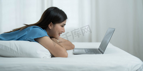 快乐、放松的年轻美丽的亚洲女性躺在卧室的床上，在笔记本上看电影或连续剧时，情绪愉快地微笑着。