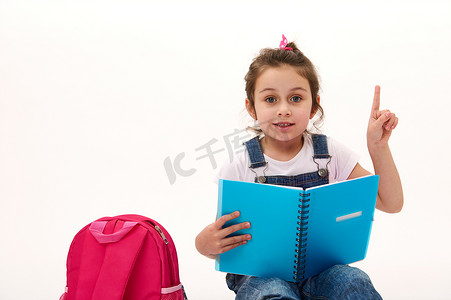 儿童教育文案摄影照片_带着文案和书包的可爱小女孩，看着相机，用食指指着白色背景上的复印空间