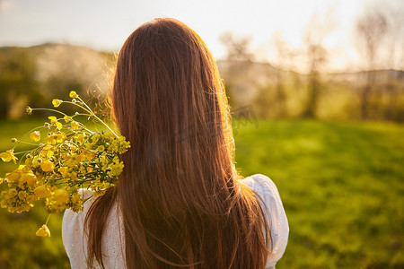 从一位红发女子的背后拍摄的照片，在夕阳的光线下拿着一束鲜花