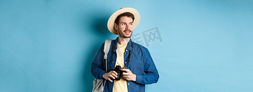 夏日拍照摄影照片_戴着夏日帽子的帅气正男，拿着相机，望向一边，游客在度假时拍照，背着背包站在蓝色背景上