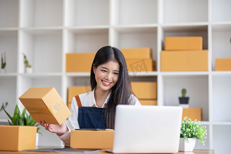 一位年轻亚洲女性的肖像，电子商务员工坐在办公室里，背景是满满的包裹，为中小企业电子商务和送货业务写下订单记录和计算器