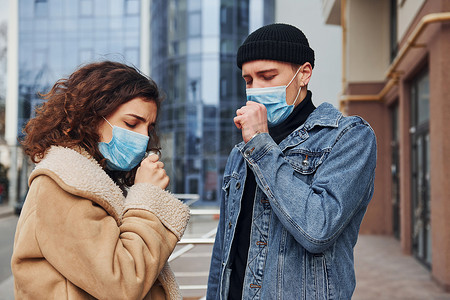 一对戴着防护面具的夫妇在检疫时间在商业大楼附近的城市户外咳嗽。