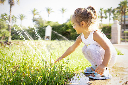 金发美女可爱的孩子弯下腰在公园喷泉的边缘，用手触摸喷水 — 概念：水对地球未来和我们孩子的重要性和价值