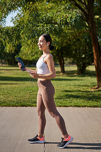 走路健身摄影照片_活跃的亚洲女孩，穿着健身服，在公园锻炼，穿着带智能手机和水瓶的运动服走路