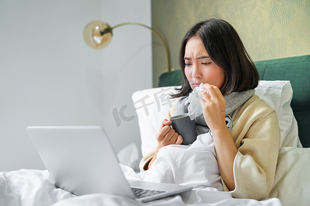 亚洲女孩打喷嚏、擦流鼻涕、感觉恶心、因流感躺在床上、看着笔记本电脑的画像