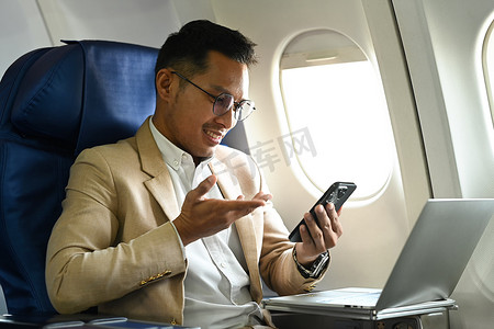 穿着正装的英俊商人坐在靠近窗户的机舱里使用手机