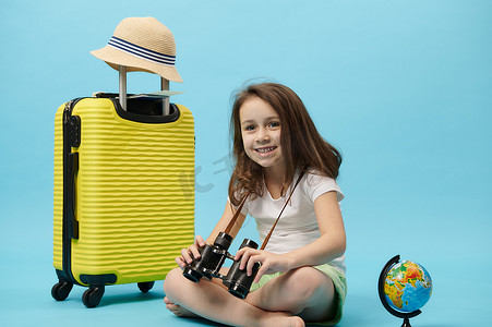 带双筒望远镜、黄色手提箱和地球仪的可爱微笑小女孩，在蓝色背景中突显