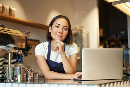 她自己摄影照片_微笑的韩国女性的画像，咖啡店的咖啡师，拿着笔记本电脑站在柜台前，在她自己的咖啡店里微笑着，看起来自信、个体经营的女企业家