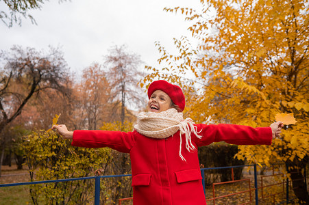 大气枫叶摄影照片_秋天，穿着红色外套和贝雷帽的快乐白人女孩手里拿着枫叶在公园里散步。