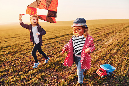 阳光明媚的白天，两个小女孩朋友在田野上玩风筝和玩具车