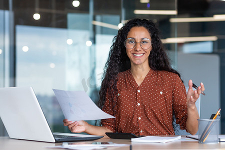 快乐而成功的西班牙裔女性的肖像，女商人微笑着看着拿着合同和发票的相机，在办公室里用笔记本电脑做纸质工作