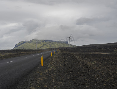 穿过冰岛北部空旷火山景观的柏油路，风力涡轮机和云层覆盖的绿色山丘，喜怒无常的天空背景，复制空间
