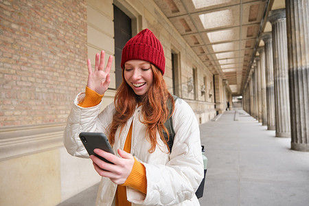 微笑的红发女孩，学生打招呼，对着智能手机摄像头挥手，在手机应用程序上与朋友视频聊天，在城市里散步