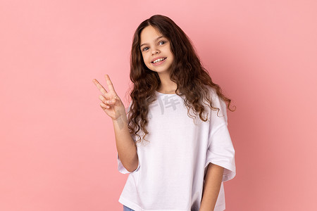 积极乐观的小女孩带着露齿的微笑看着镜头，展示 v 形手势，和平手势。