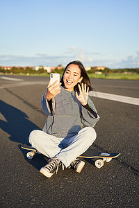 快乐的亚洲少女坐在滑板上自拍，在智能手机应用程序上进行视频聊天，溜冰者坐在长板上录制视频博客