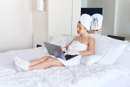 双11贴片摄影照片_淋浴后，一个裹着毛巾的女孩在笔记本电脑上工作，并在眼睛、嘴唇和手套下方的皮肤上贴上化妆品贴片，以滋润她的手脚。