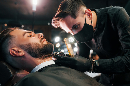 发型时髦的年轻人坐着，在理发店被戴黑色防护面具的男人剃胡子
