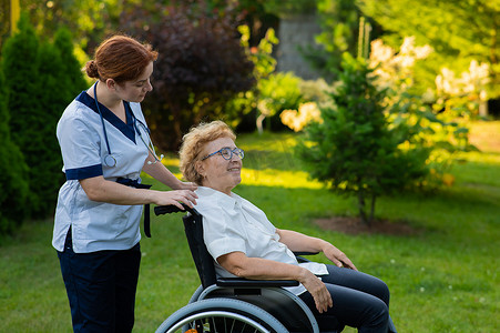 白人女医生和一位坐在轮椅上的老年病人在公园里散步。