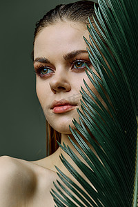 修饰背景摄影照片_一位化着晚妆的精致、优雅的女士站着，一片绿色的棕榈叶遮住了她的一部分脸。