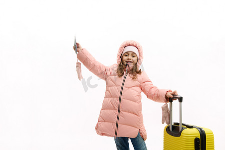 可爱的小女孩穿着温暖的粉色冬衣，带着机票、登机牌和黄色手提箱，挥舞着你好相机